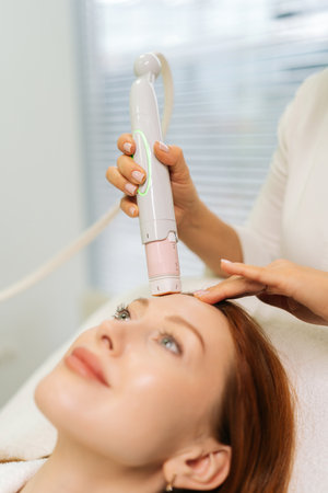Close-up vertical shot of dermatologist using modern device performing mesotherapy procedure on forehead of woman lying on treatment table in beauty salon. Concept of anti-age rejuvenation skincareの写真素材