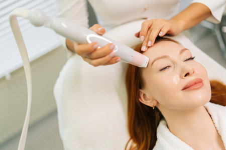 Close-up cropped shot of unrecognizable beautician using modern device performing mesotherapy procedure on forehead of young woman lying on treatment table in modern beauty salon.の写真素材