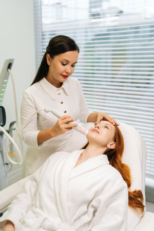 Vertical shot of beautiful young woman receiving microneedling rejuvenation treatment in clinic of aesthetic medicine. Cosmetologist performing needle mesotherapy on female client face.の写真素材