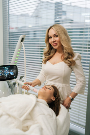 Vertical portrait of beautiful beautician performing mesotherapy treatment with specialized device on woman facial skin looking at camera, targeting skin rejuvenation and age-defying results.の写真素材