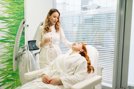 Skilled cosmetic professional doctor in white uniform discussing skin treatment options with female client lying on couch in modern beauty salon. Concept of non-surgery cosmetology, skincare.の写真素材
