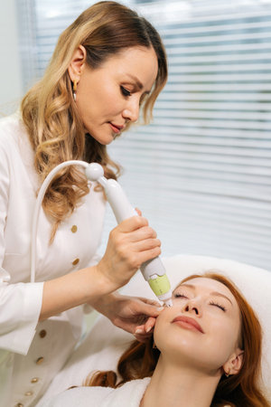 Process of skin rejuvenation procedure from of microneedling professional performing on female lying with closed eyes on treatment bed, targeting dermal renewal with precision handheld deviceの写真素材