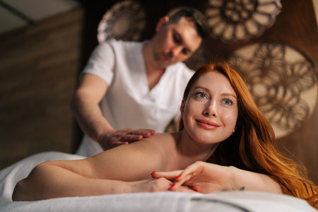 Portrait of happy smiling redhead woman lying on massage table, enjoying relaxing back massage by professional masseur male in spa environment with decorative interior on background.の写真素材