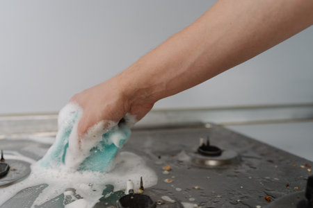 Housewife hand wiping gas stove with sponge and detergent, promoting hygiene and achieving sparkling clean kitchen surface removing dirt and grease. Concept of domestic work, housekeeping, household.の写真素材