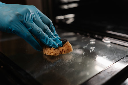 Detail cropped shot of unrecognizable man wearing blue gloves wiping glass door of oven with sponge, ensuring thorough cleaning and maintenance of kitchen appliances. Concept of hygiene, cleanliness.の写真素材
