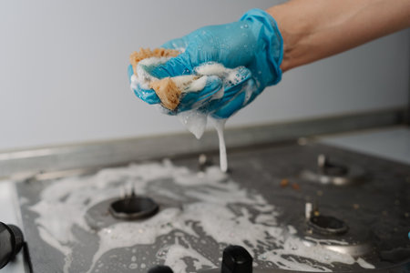 Close-up hands of unrecognizable man in gloves squeezing soapy water from sponge during cleaning dirty gas stove, to removing stubborn grime and grease. Concept of domestic work and household.の写真素材
