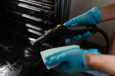 Cleaning person wearing blue gloves using steam cleaner and sponge to clean interior of dirty gas oven, ensuring hygiene and maintenance, close-up. Concept of maintenance modern home appliance.の写真素材