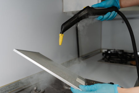 Close-up of professional cleaning technician wearing protective gloves sanitizing kitchen extractor filter using high-pressure steam cleaning equipment over gas stove. Concept of hygiene, cleanliness.の写真素材