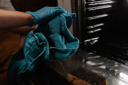 Professional cleaner wearing blue gloves wiping hands with turquoise towel after cleaning modern kitchen oven, ensuring hygiene and cleanliness. Concept of domestic work, housekeeping and household.の写真素材