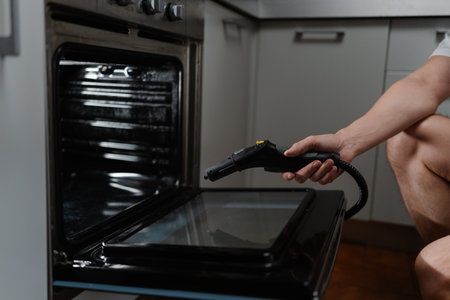Side view cropped shot of unrecognizable housekeeper steam cleaning kitchen oven surface, targeting stubborn grease with powerful cleaning equipment for spotless, sanitized cooking space, close-up.の写真素材