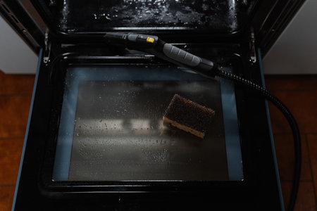 Top view of steam cleaner and sponge resting on inner glass of gas oven door during cleaning process, with glistening water droplets accentuating surface, close-up, nobody, no people.の写真素材