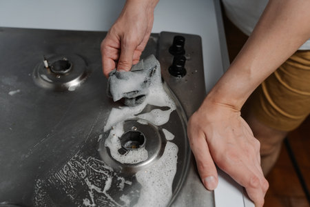 Close-up top view of housekeeper male hands scrubbing gas stove with detergent and rag during hygiene and maintenance in kitchen environments. Concept of domestic work, housekeeping and household.の写真素材
