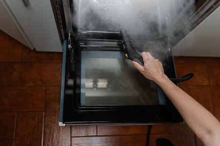 High-angle view of cleaning company worker washing gas oven with steam cleaner in kitchen, holding steam cleaner gun removing grease and ensuring hygienic cooking environment, close-up cropped shotの写真素材