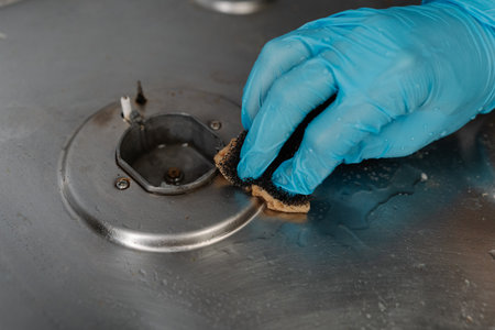 Housekeeper hand wearing blue glove cleaning dirty gas stove, removing grease and food remnants with sponge and detergent, close-up. Concept of household chores and kitchen hygieneの写真素材