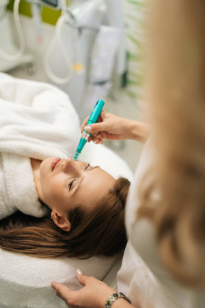 Close-up vertical shot of cosmetic professional doctor performing microneedling treatment on female client face for skin rejuvenation and collagen stimulation, using modern device in beauty clinic.の写真素材