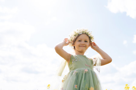 Shooting from below of cute preschooler girl wearing flower crown and fairy green dress, enjoying sunny summer day in meadow, looing at camera with cheerful expression. Concept of happy childhood.の写真素材