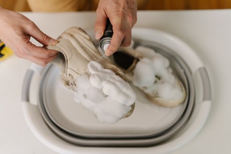 Male hands applying foam cleaner on white sneakers in basin, detailing shoe cleaning technique and maintenance for pristine footwear condition, close-up. Concept of shoe care and maintenance practicesの写真素材