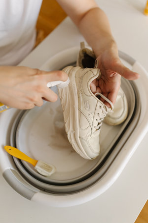 Vertical top view of person carefully cleaning dirty white sneakers using brush, detergent and small portable basin, ensuring shoes thoroughly cleaned and refreshed, close-up.の写真素材