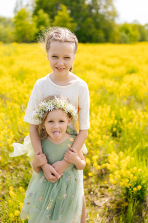 Siblings hugging in sunlit wildflower meadow, younger sister wearing floral crown playfully teasing with tongue out. Two sisters embracing standing posing in green meadow on sunny summer day.の写真素材
