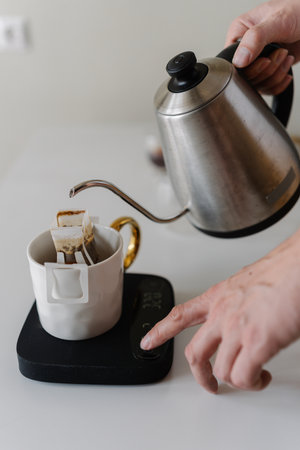 Male hands adjusting kitchen scales during preparing drip coffee, pouring hot water from kettle onto coffee bag on kitchen scales in precise brewing process, close-up, creating morning ritual.の写真素材