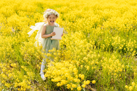 Cheerful child girl with flower crown fooling around and showing tongue looking at camera, holding blank white canvas in vibrant yellow field, offering picturesque setting for mockup designs.の写真素材