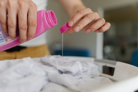 Housekeeper male pouring stain remover from pink bottle onto white clothing soaking in plastic basin, treating fabric before laundry washing cycle, close-up. Concept of household cleanliness.の写真素材