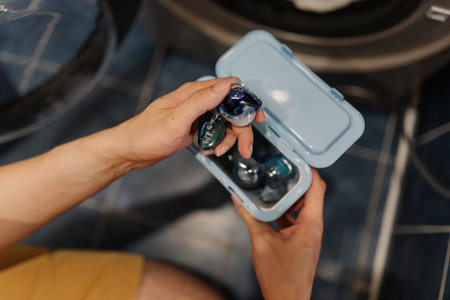 Top view of unrecognizable man holding two laundry detergent pods preparing to load washing machine with clothes for fresh and clean laundry. Concept of household chores and modern appliance usage.の写真素材
