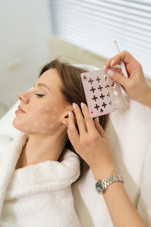 Vertical cropped shot of beautician preparing patient facial skin for treatment marking grid with white pencil in modern beauty clinic. Cosmetologist applying pre-procedure markings on woman face.の写真素材