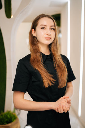 Professional female cosmetologist wearing black uniform looking at camera, standing confidently with crossed hands in wellness center, representing skilled beauty industry expert.の写真素材