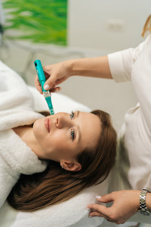 Vertical shot of cosmetic professional doctor performing microneedling treatment on female client face for skin rejuvenation and collagen stimulation, using modern device in beauty clinic.の写真素材