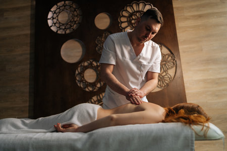 Shooting from below of massage therapist providing relaxing back massage to redhead woman lying on massage table, enjoying spa treatment in wellness center. Concept of healthy lifestyle and wellbeing.の写真素材