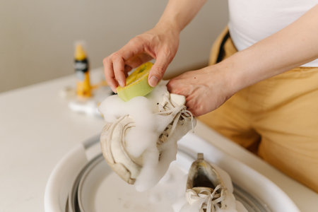 Man cleaning white sneakers with foam and brush, scrubbing dirty athletic footwear in household basin, performing domestic cleaning task, close-up. Concept of professional footwear maintenance.の写真素材