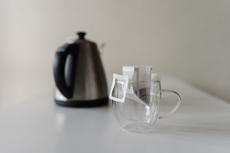 Close-up of drip coffee bag placed in glass cup, ready to brewed with hot water from modern electric kettle placing on white background. Concept of convenient, quick way to enjoy fresh cup of coffeeの写真素材