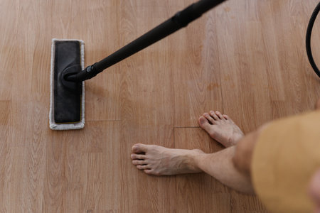 Top view of barefoot male cleaning hardwood floor with steam mop during house cleaning, close-up. Maintaining hygiene and cleanliness in home environment. Concept of cleaning and household.の写真素材