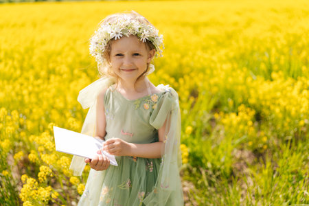 Portrait of cute child girl wearing green fairy dress holding blank white paper in vibrant yellow field, offering picturesque setting for mockup designs. Concept of happy childhood.の写真素材