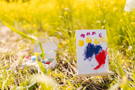 Vibrant finger painting, child artwork, displayed on small easel in green meadow full of yellow flowers on sunny summer day, with drawing set nearby, evokes sense of outdoor creativity and joy.の写真素材