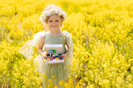 Portrait of cute child girl in fairy green dress holding box of watercolors, ready to capture beauty standing in green meadow full of yellow flowers on bright sunny summer day, smiling looking camera.の写真素材
