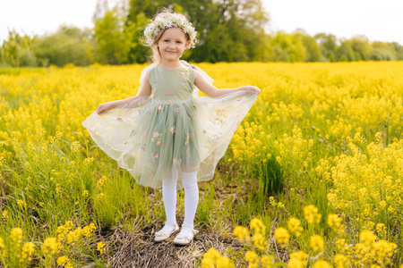 Charming young girl adorned with flower crown, twirling in sunlit meadow surrounded by golden wildflowers, radiating pure childhood happiness, enjoying beautiful nature and smiling looking at camera.の写真素材