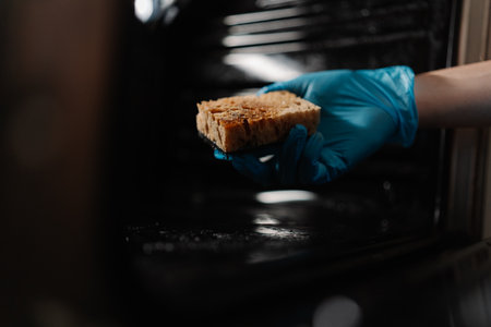 Cropped shot of cleaning company worker in blue rubber glove holding dirty sponge, scrubbing soiled gas oven covered in grease stains and splashes, tackling mess in kitchen environment, closeup.の写真素材
