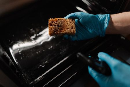Top view of cleaning company worker in gloves spraying detergent on sponge, preparing to clean glass door of modern oven in kitchen with dark interior. Concept of cleanliness during routine chores.の写真素材