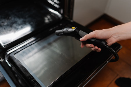 Close-up cropped shot of unrecognizable man steam cleaning kitchen oven surface, targeting stubborn grease with powerful cleaning equipment for spotless, sanitized cooking space at home.の写真素材