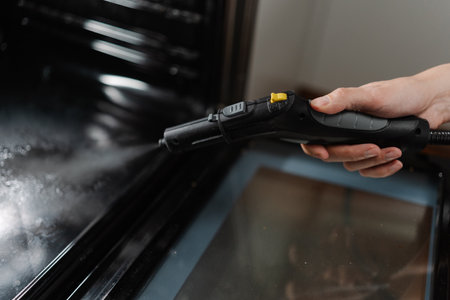 Professional cleaning service employee male cleaning oven with steam cleaner in kitchen, holding steam cleaner gun removing grease and ensuring hygienic cooking environment, close-up cropped shotの写真素材