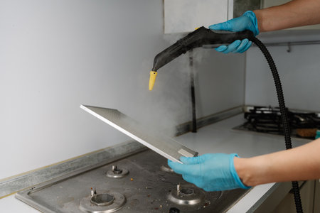 Cleaning company worker male wearing protective gloves sanitizing kitchen extractor filter using high-pressure steam cleaning equipment over gas stove. Concept of maintenance modern home appliance.の写真素材