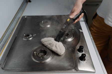 Top view of housewife cleaning dirty gas stove using steam cleaner and cloth, removing grease and burnt food residues, ensuring hygienic kitchen environment. Concept of modern housekeeping appliance.の写真素材