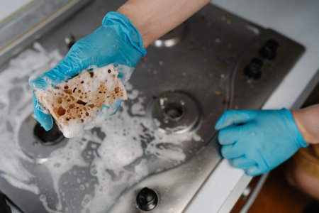 High-angle view of housewife in protective gloves squeezing soapy water from sponge during cleaning dirty gas stove, to removing stubborn grime and grease. Concept of domestic work and household.の写真素材