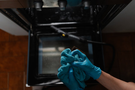 High-angle view of housekeeper male wearing blue gloves squeezing out wet rag after cleaning inside of gas oven with steam cleaner on blurred background. Concept of housekeeping and cleaning.の写真素材
