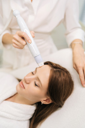 Vertical shot of beautiful female patient having microneedling rejuvenation treatment in clinic of aesthetic medicine. Beautician using device to performing mesotherapy treatment to woman foreheadの写真素材