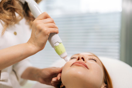 Close-up of aesthetician using microneedling device performing rejuvenation procedure to female patient lying with closed eyes on treatment table, enhancing skin health and appearance in spa salon.の写真素材