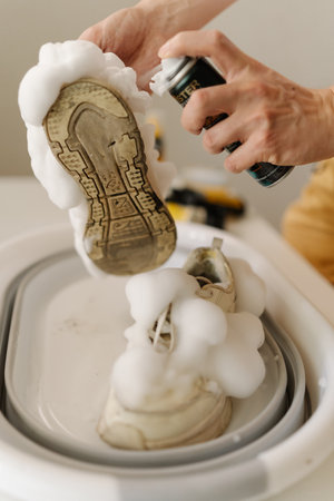 Close-up of young man applying foam cleaner on dirty white sneakers in basin, highlighting shoe cleaning process and emphasizing proper care. Concept of shoe maintenance and hygiene.の写真素材