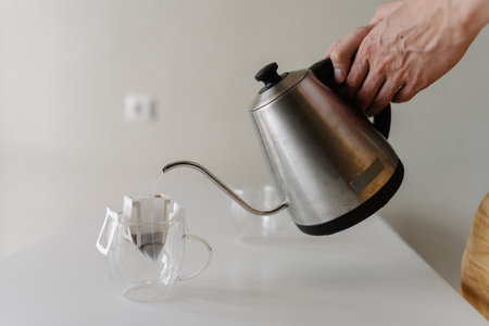 Close-up of unrecognizable young man preparing drip coffee bag by pouring hot water from stainless steel kettle into clear glass cup placing on white table at home, creating inviting morning ritual.の写真素材